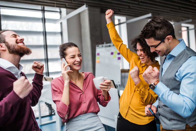 A group of young businesspeople with smartphone standing in office, expressing excitement.